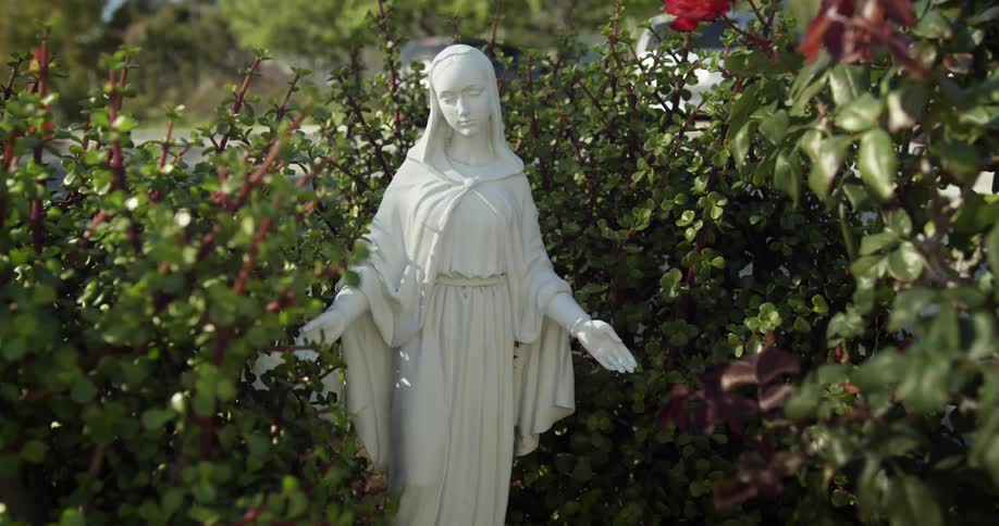 A white statue of the virgin mary with arms outstretched stands in a garden