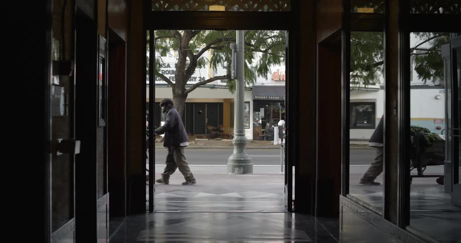From the inside of a building a homeless man pushing a shopping cart walks by the entrance