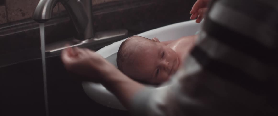 A woman giving a baby a bath by a sink with water coming out of the faucet