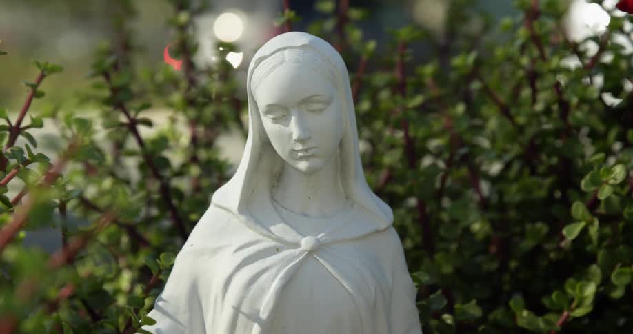 A statue of a woman wearing a hooded scarf sits in front of a dense bush outside