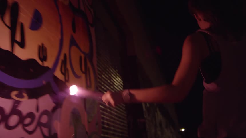 A woman with a flare skipping by a graffiti covered wall at night