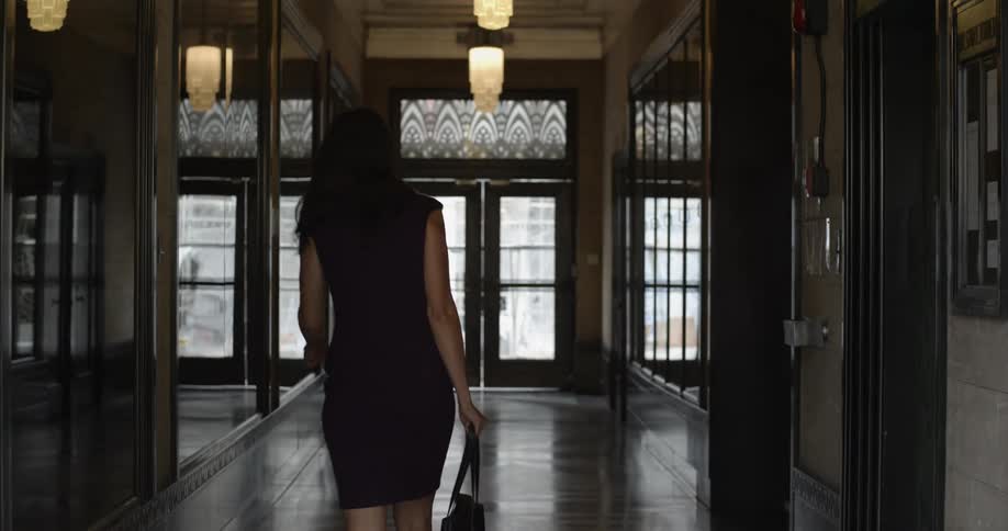 A woman walks down a corridor and through a doorway while carrying a purse
