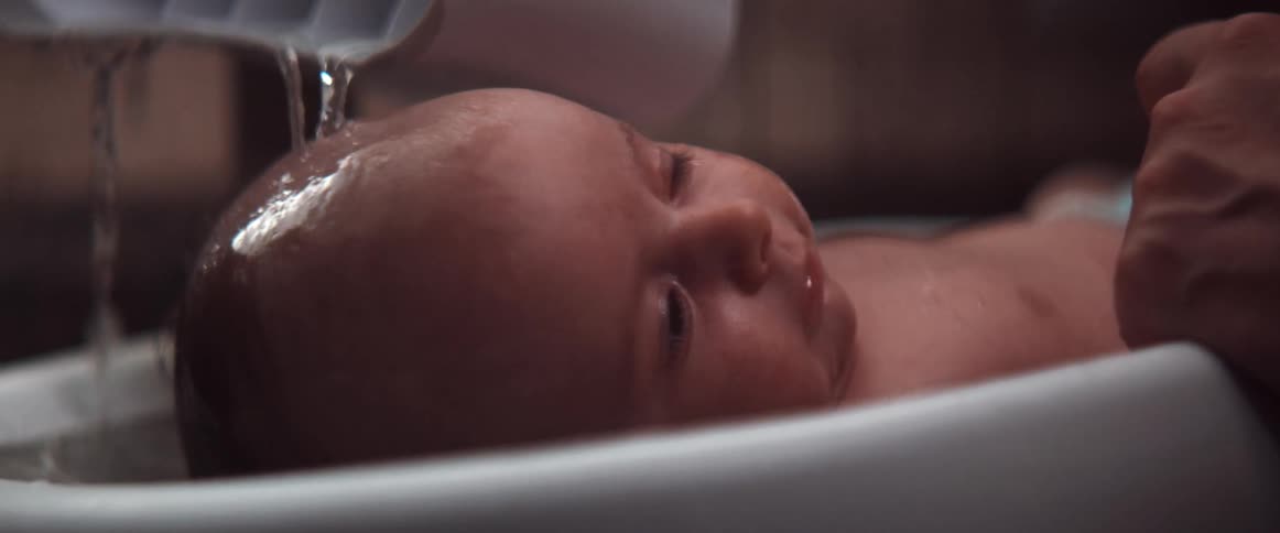 A parent pours water from a cup onto a babies head in a bathtub