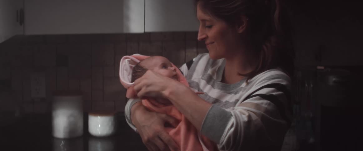 A mother smiling while holding her swaddled newborn in a kitchen