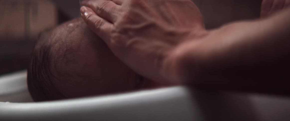A woman covers a babys eyes with her hand before pouring water from a jug over his head at bath time