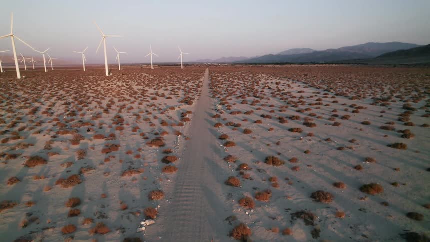 A group of wind turbines in the desert are spinning next to a long dirt road