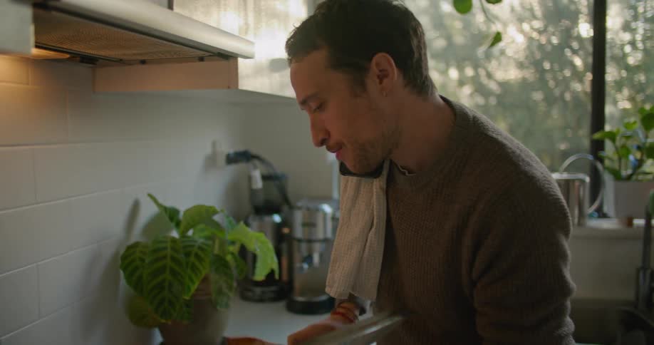 A man cooking at home tastes the dish as he talks on his cell phone