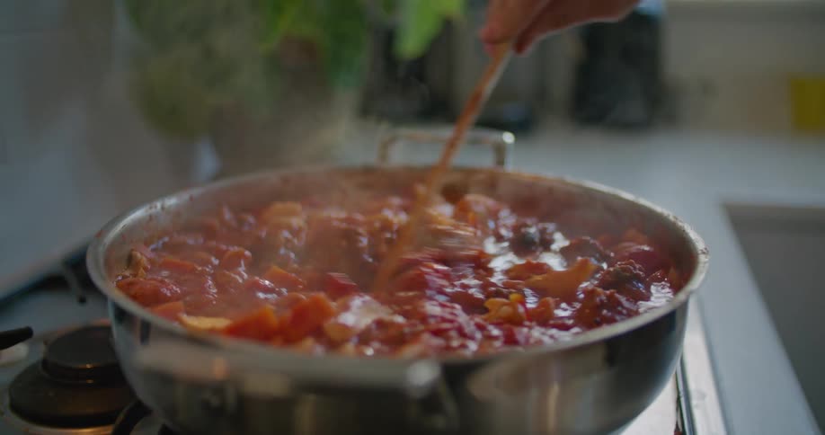 A man takes the lid of a skillet and stirs a tomato based stew
