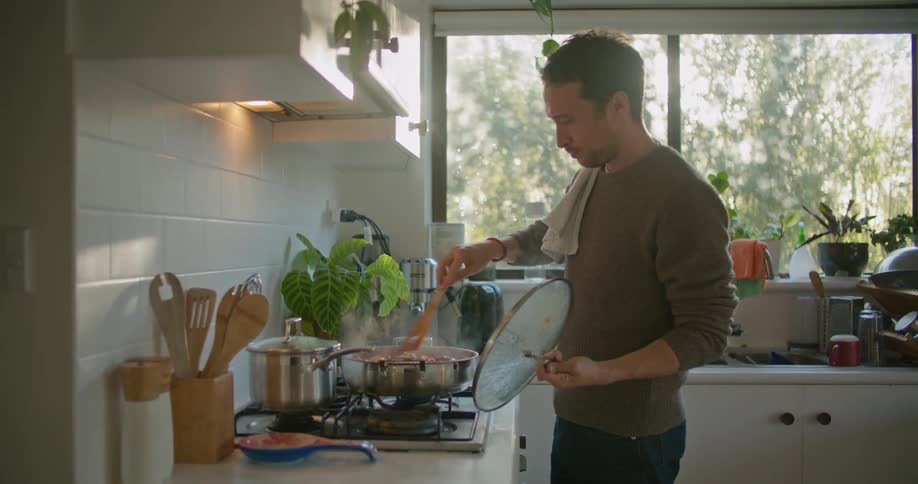 A man in a home kitchen walks to the stove and tastes what is cooking on the stove