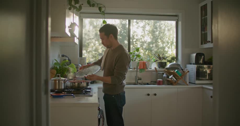 A man cooking at a stove opens the lid of a pan and tastes what is inside
