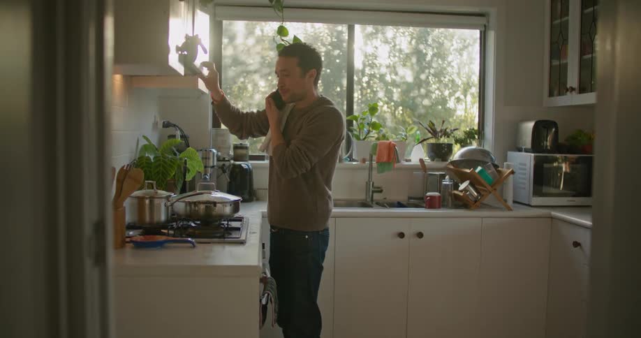 A man smiles and talks on the phone while stirring and tasting food on the stove