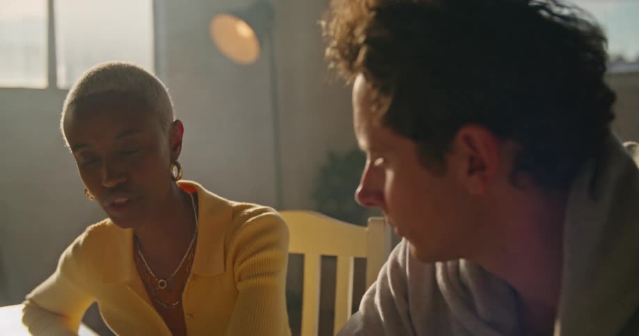 Woman with short hair hoop earrings and yellow sweater and man with gray hoodie talk with one another in office