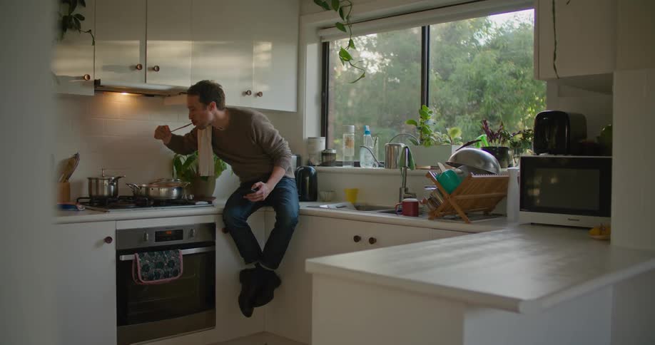 A man in a kitchen using a smart phone while sitting on the countertop and cooking on the stove