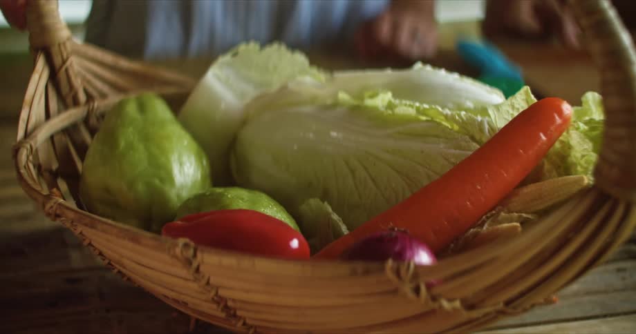 Woman with white and blue dress takes carrot out of vegetable basket to cut on cutting board
