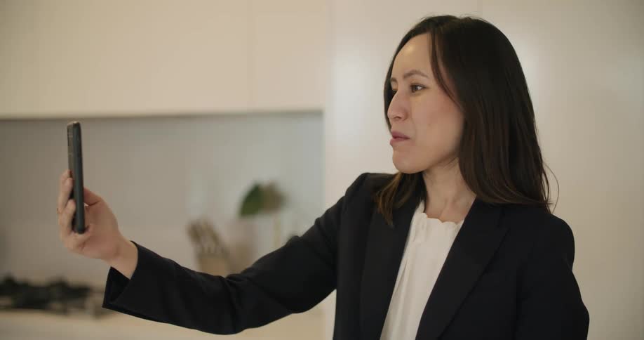 A woman holds her smartphone out in front of her face during a video call in her kitchen