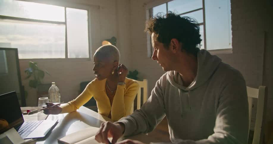 Man and woman sit at table in office with laptops discussing screen contents