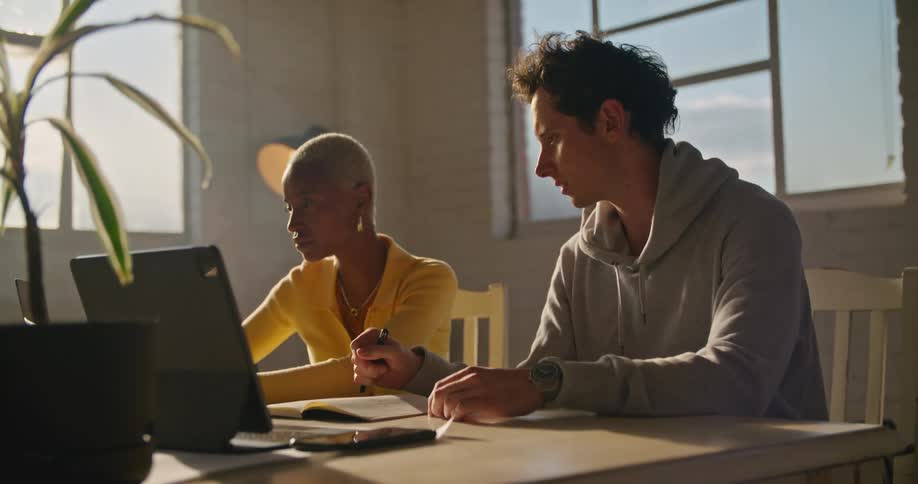 Woman with short hair hoop earrings and yellow sweater and man in gray hoodie sits at table working on laptop in office