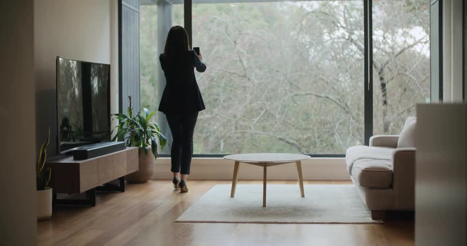 Woman walks around living room and toward large window showing leafless trees as she uses smartphone to make video