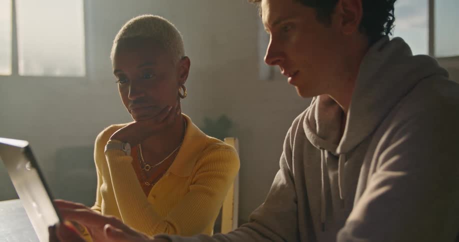 A woman and a man are sitting and talking as they work on a laptop