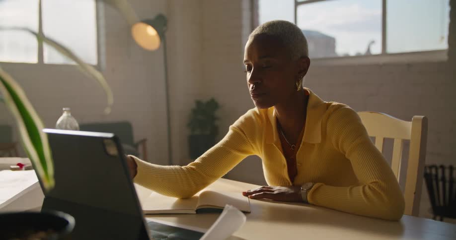 A woman is sitting inside with a pencil in hand and talking while pointing at a laptop