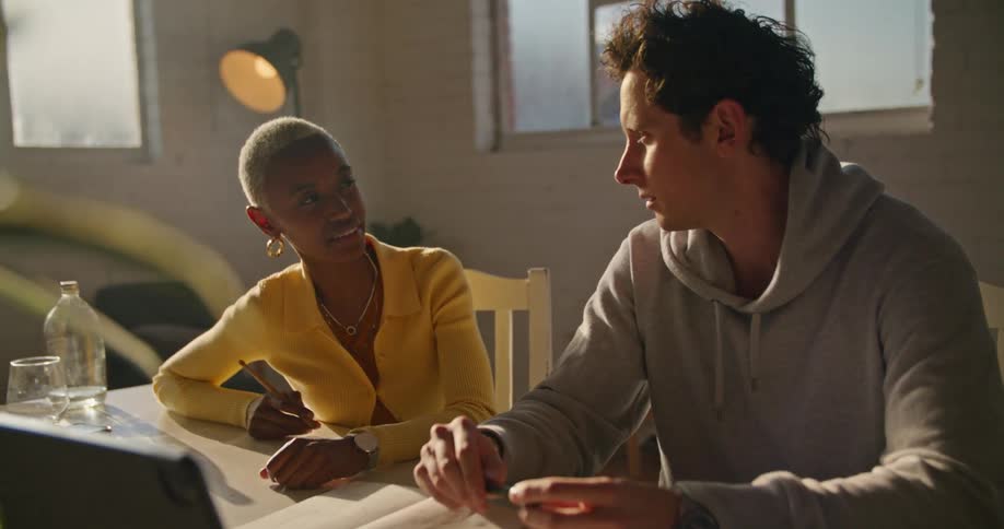 A man and woman sitting side by side at a table talking with a notebook open and phone and computer at hand