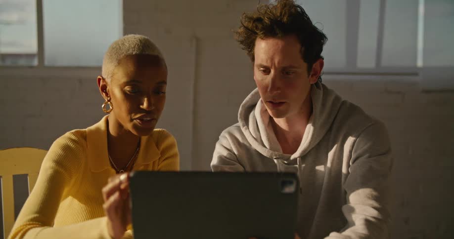 A man and a woman in a room looking at a laptop with a wall and windows in the background