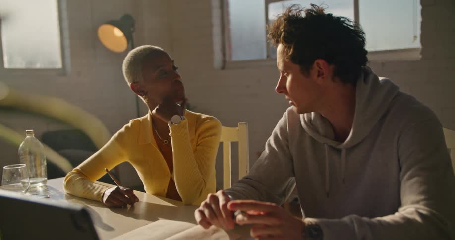Woman with short hair hoop earrings and yellow sweater sits at table with man wearing grey hoodie talking together in the office