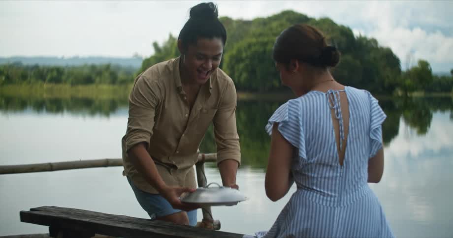 A man bringing a plate of food to a woman with a river the sky and clouds in the background