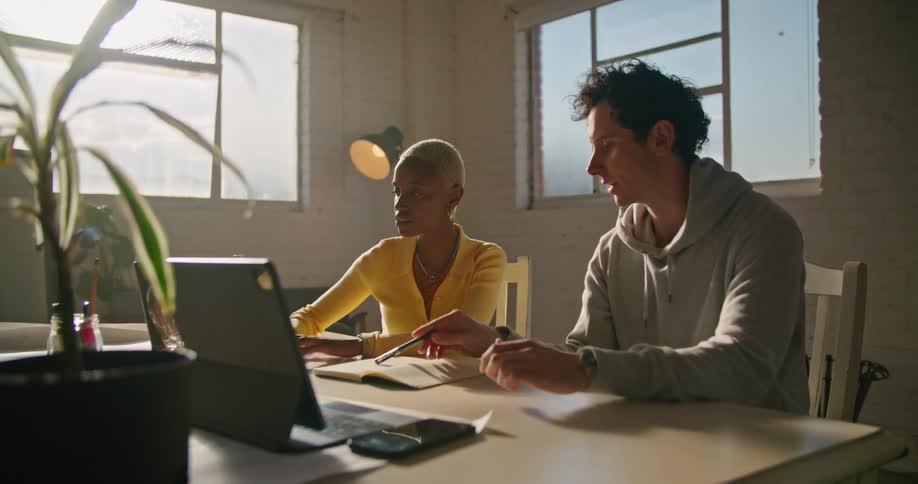 Woman with short hair hoop earrings and yellow sweater and man in gray hoodie work on laptop talking to one another