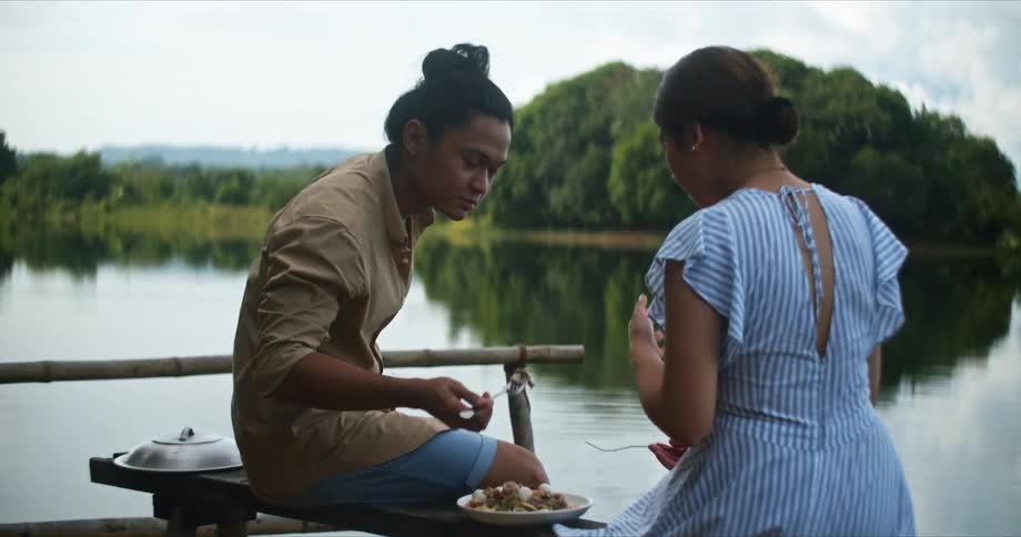 A teenage boy and girl sit on the shore with a plate of food and he picks up a forkful and offers it to the girl
