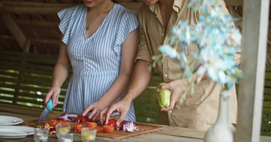 Man with long hair brown shirt and woman with white blue dress cut vegetables on cutting board