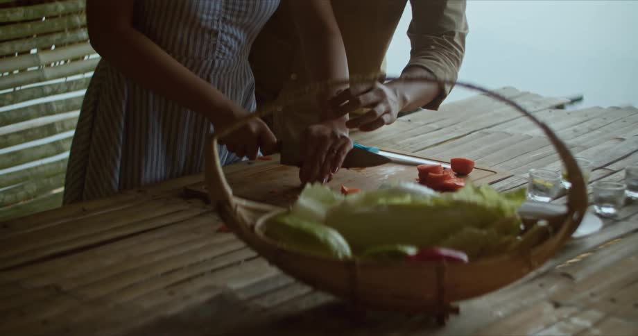 Man with long hair and brown shirt shows how to cut vegetables on cutting board to woman with white and blue dress next to lake
