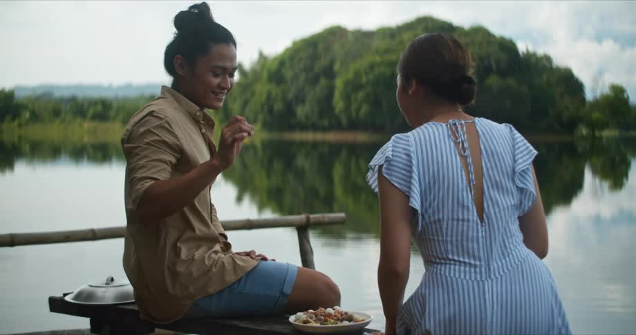 Man and woman sitting on bench prepares to eat food next to still lake