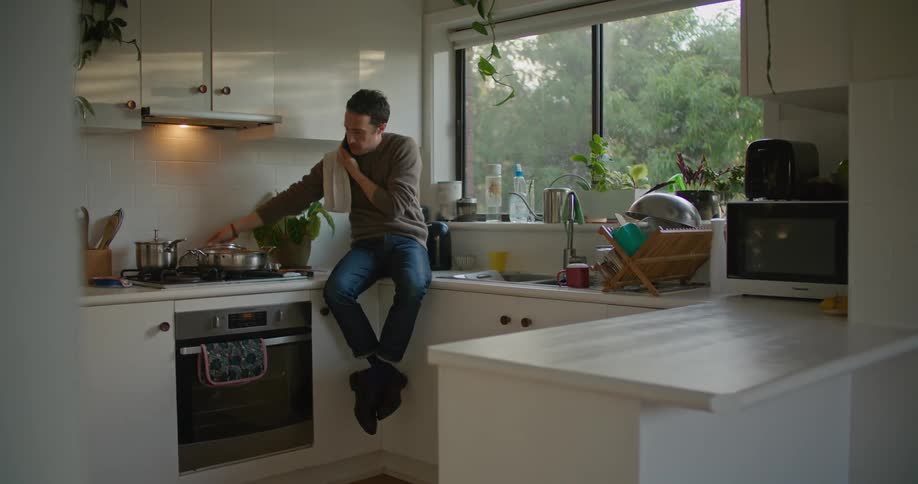 A man is sitting on the counter as he talks on the phone and stirs some food on the stove