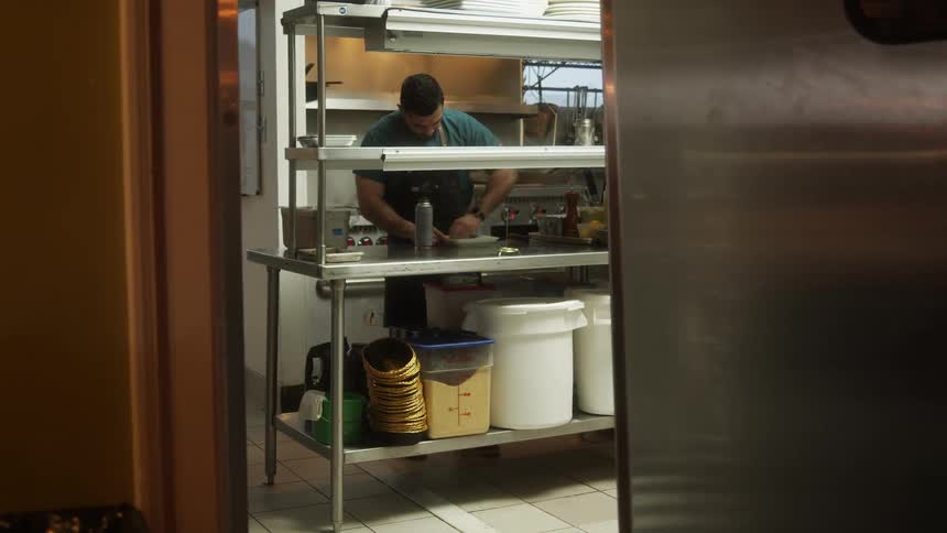 A man walks to a stainless counter in a restaurant kitchen and wipes a pan