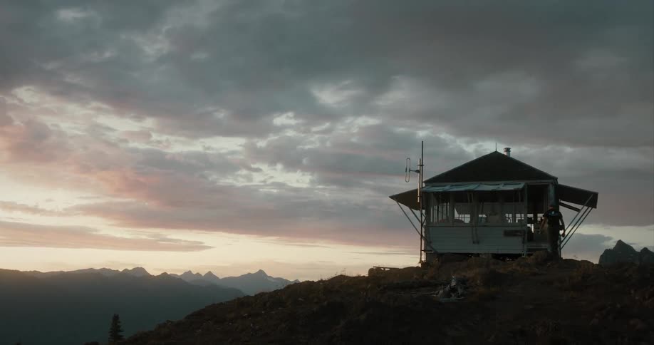 A silhouetted person walking towards a small building in mountains at sunset