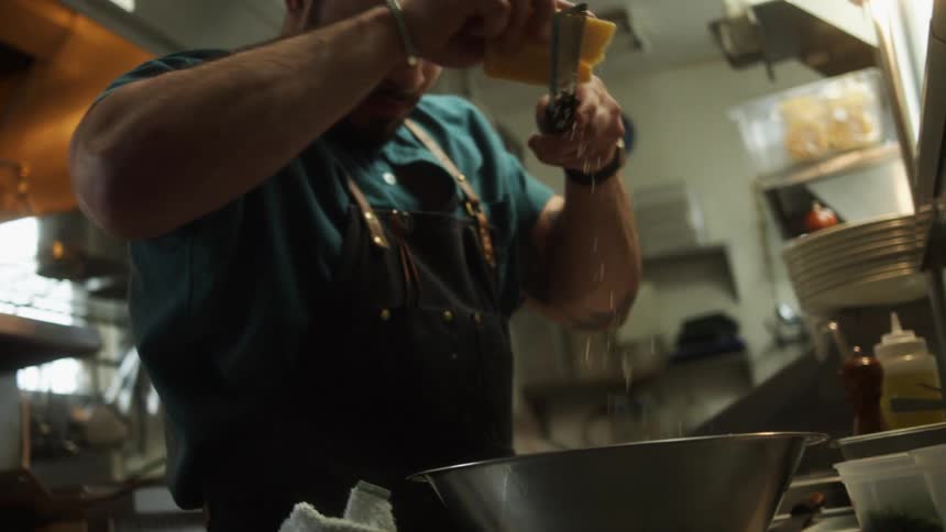 A male chef wearing an apron grates parmesan cheese into a metal bowl on a chefs table