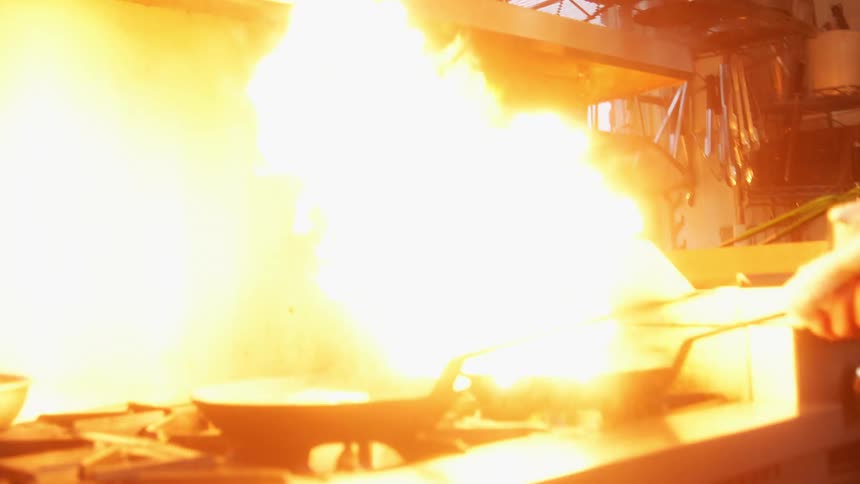 A cook pours oil into a skillet causing flames to erupt while cooking in an industrial kitchen