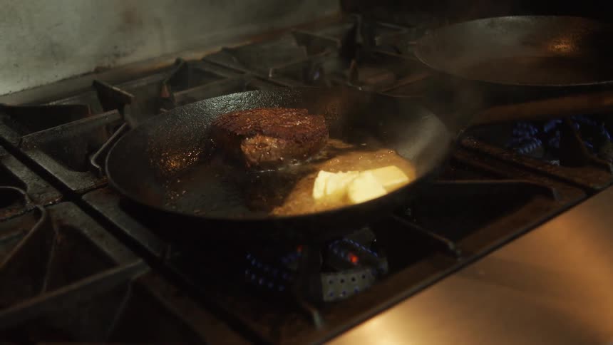 Chef frying a piece of meat with butter on a pan over the kitchen range inside a restaurant
