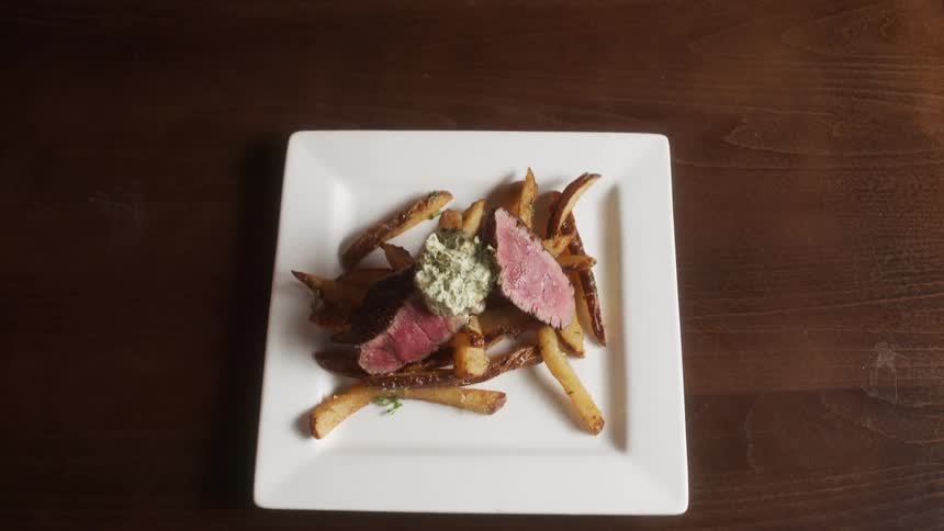 A square white plate holds rare cooked steak herb butter on top of fries