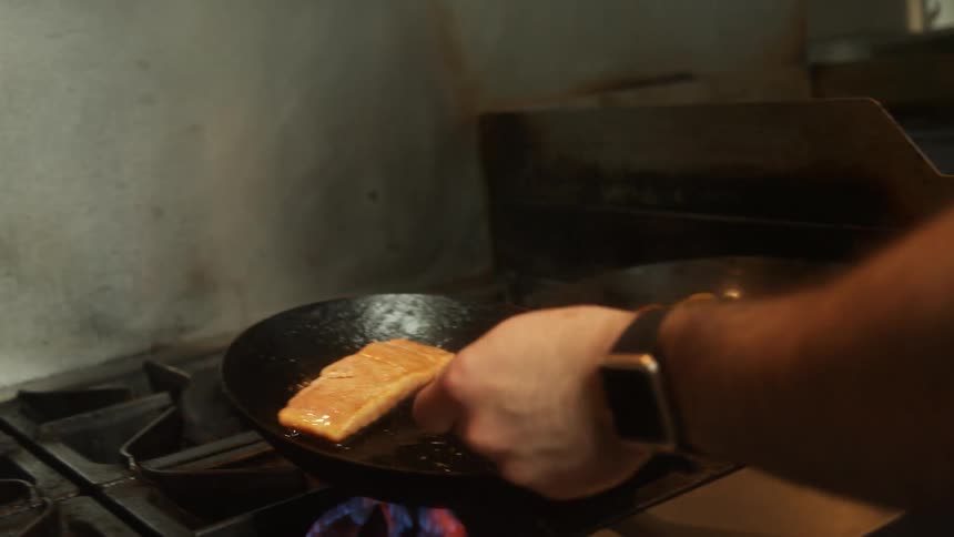A person frying a piece of fish in a frying pan and some shrimp in another frying pan on a stove