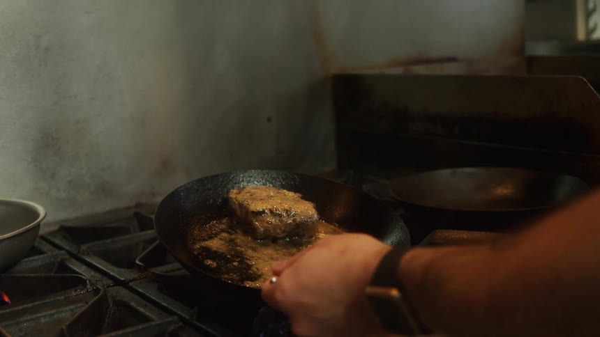 A man uses a spoon to scoop gravy over a steak frying in a skillet