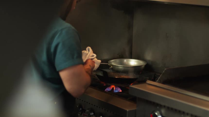 A man is tending to two skillets on a gas range