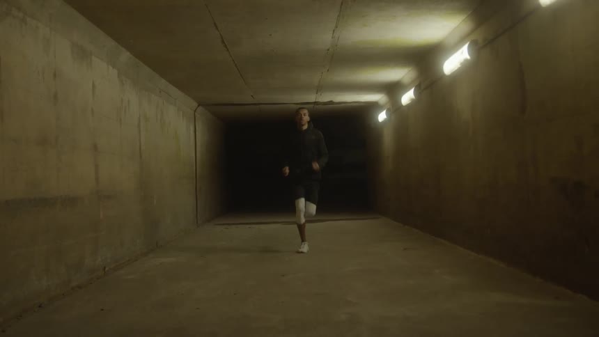 A black man jobs through a concrete tunnel lit by mounted lights