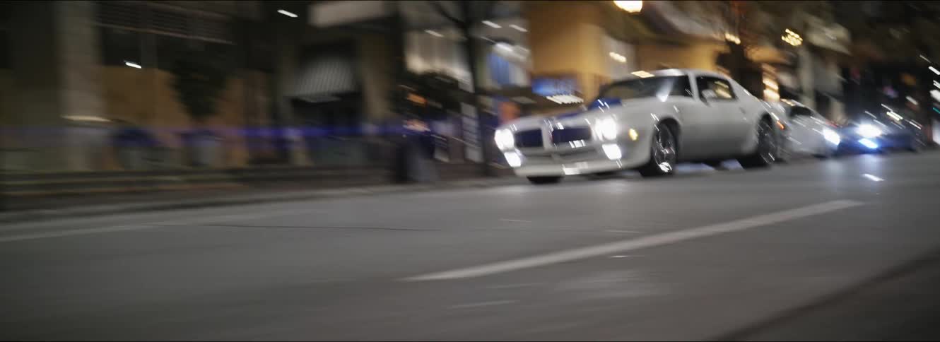 A white trans am drives on a downtown city street at night