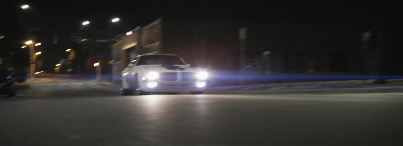 A car with headlights driving on a city street with buildings in the background