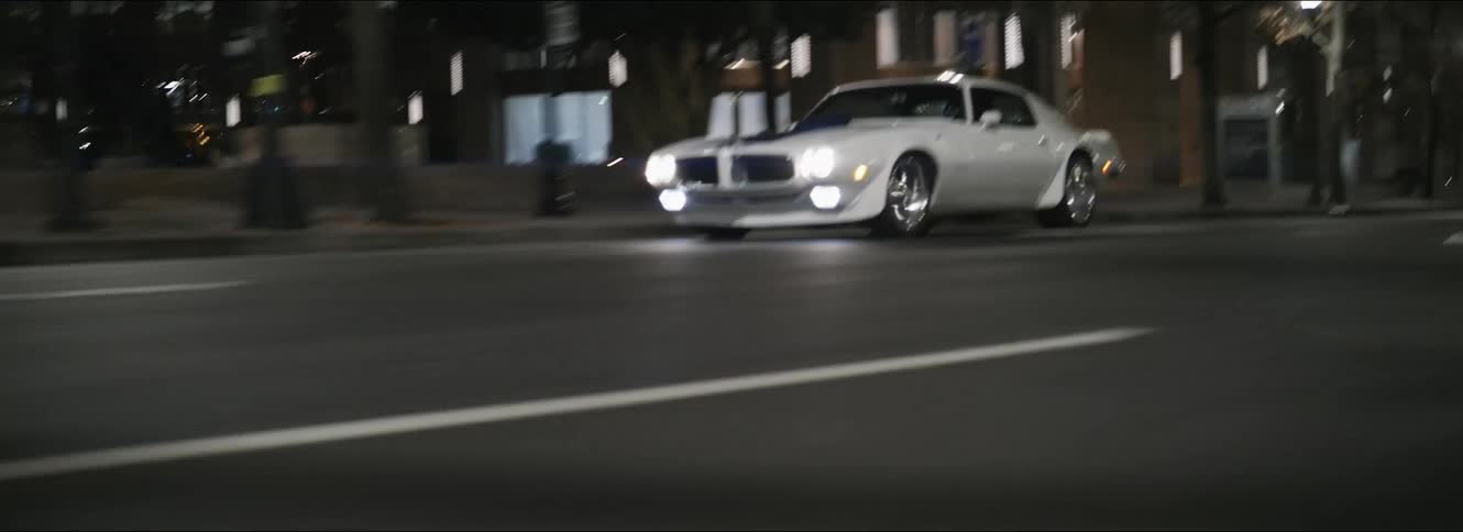 A white trans am drives on a downtown city street at night