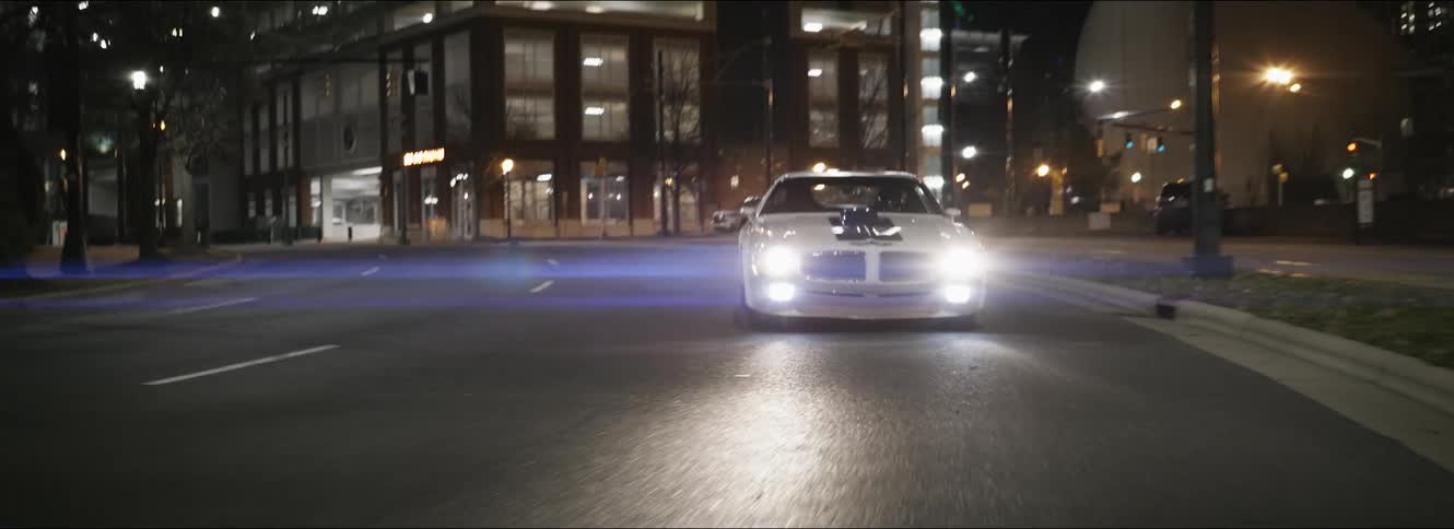 A car with headlights driving on a city street with buildings in the background