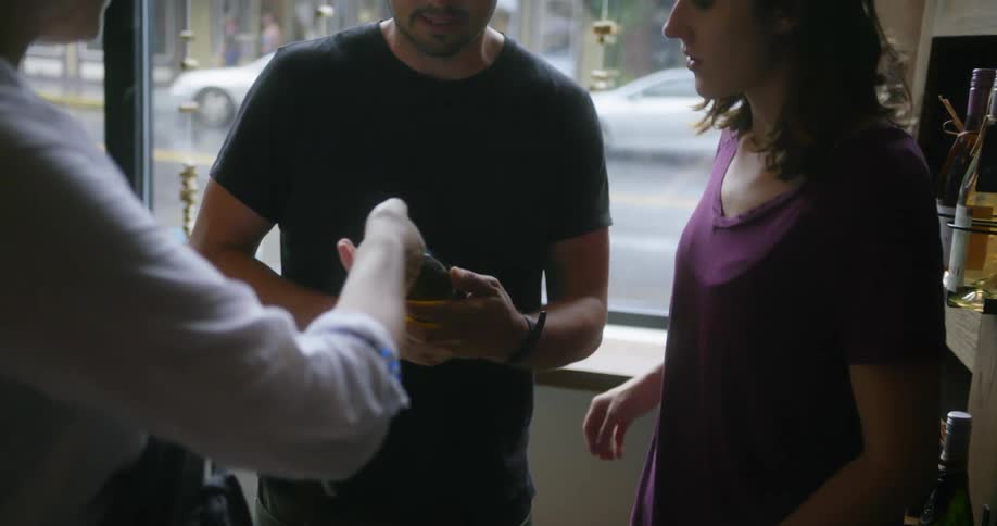 An employee takes a bottle of wine from a shelf and hands it to a customer who looks at it with his friend