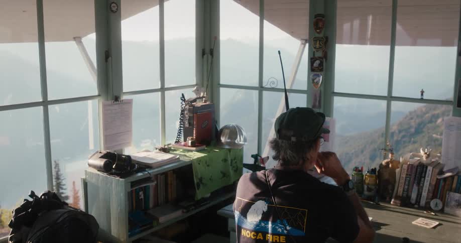 A man sits in a building on top of a hill and talks on a walkie talkie as he looks out the window